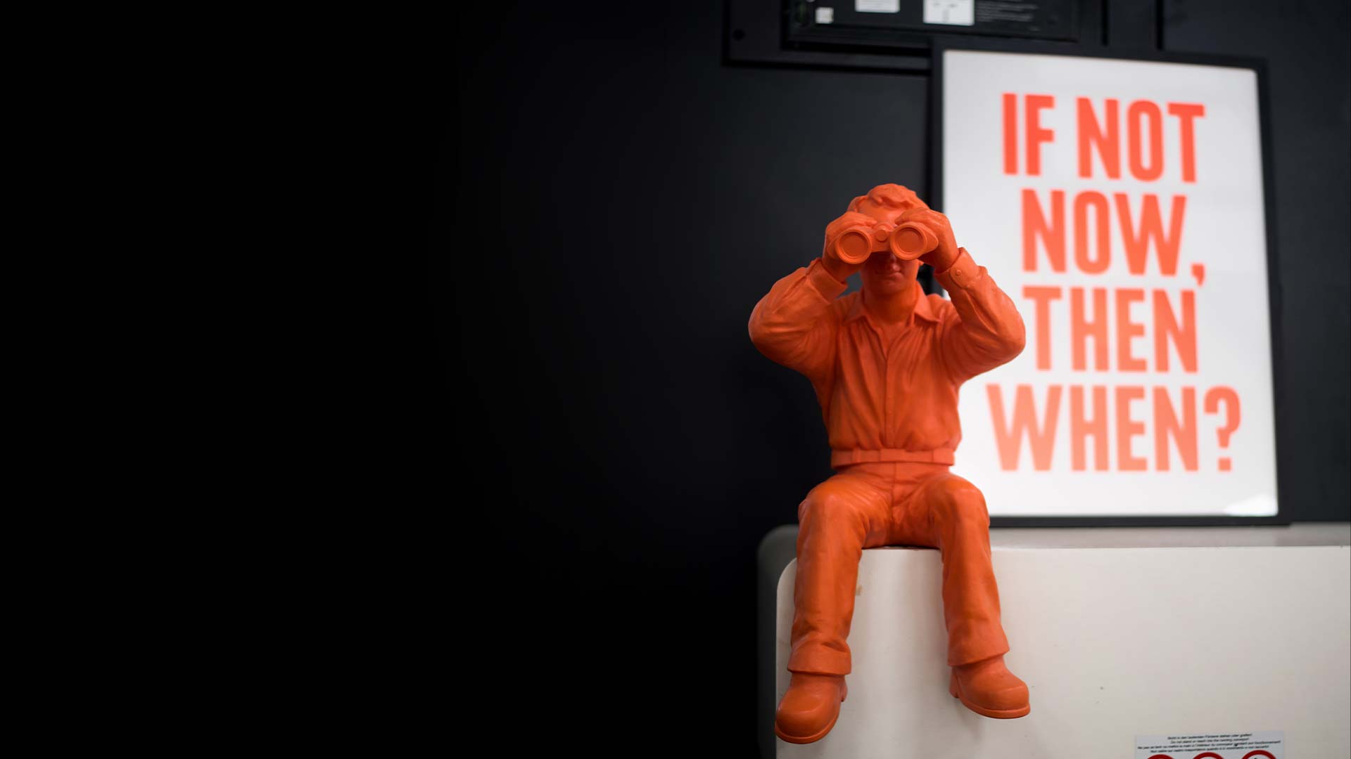 Orange Figur mit Fernglas vor dem Plakat "If not now, then when?"
